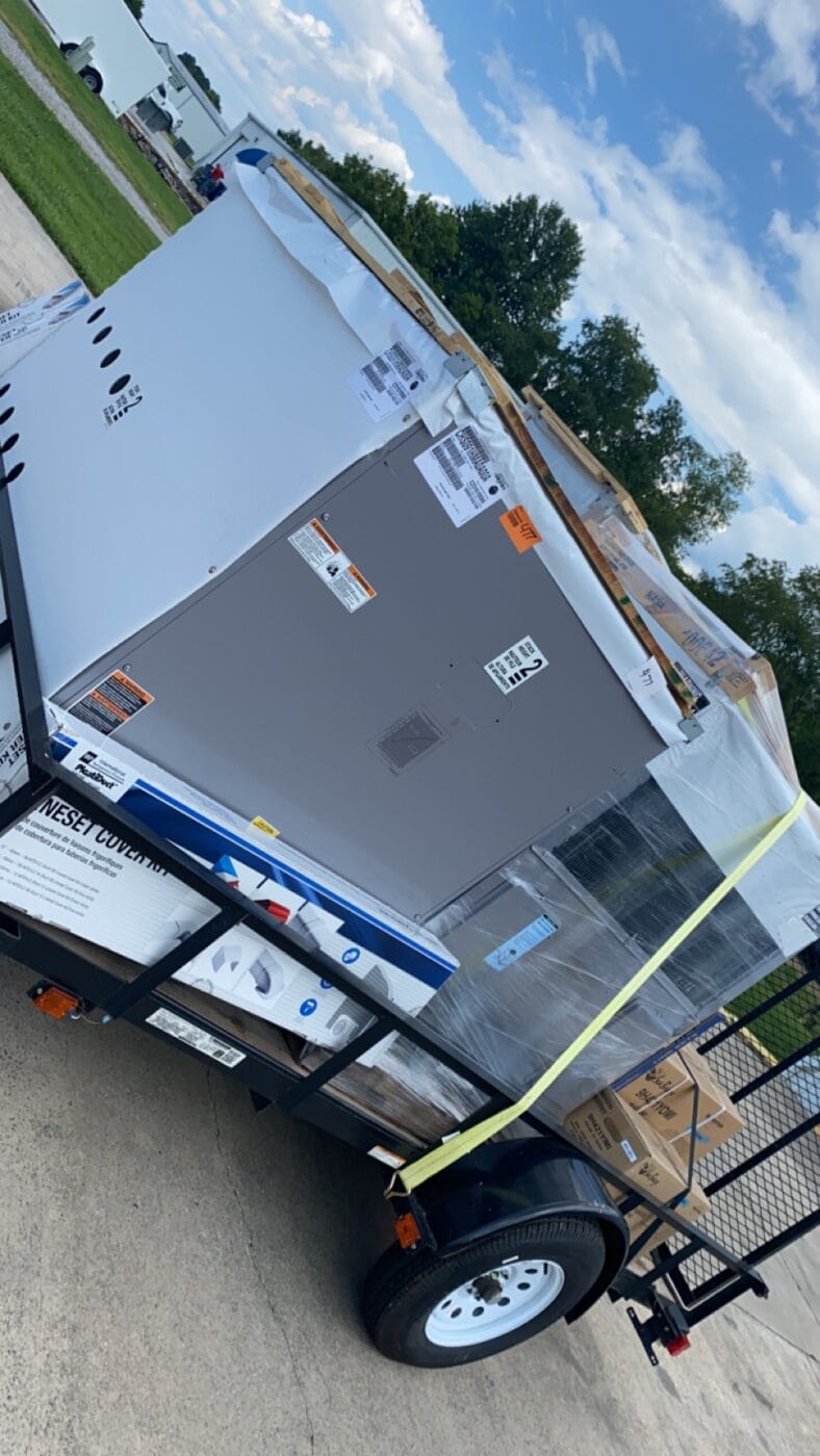 Large appliance secured on a trailer with straps, surrounded by packaging. Outdoor setting with trees and a blue sky in the background.