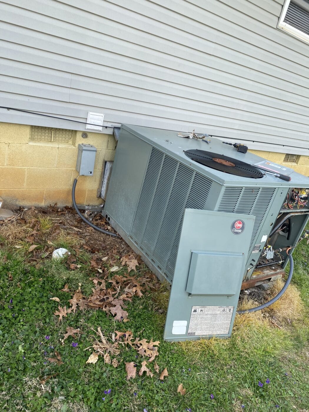 An air conditioning unit partially tipped over next to a building with visible wiring. The surrounding area has fallen leaves and patches of grass.