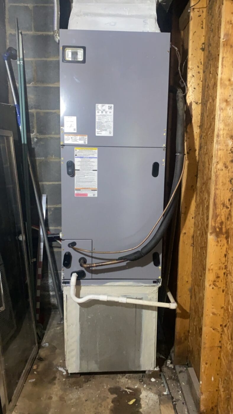 A vertical gray HVAC unit with attached ducts and pipes installed in a basement corner. Nearby walls are concrete and wood. A broom and panel are leaning against the wall.