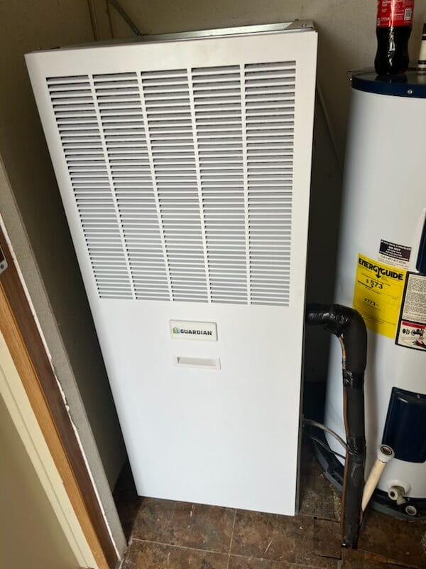 White Guardian heating unit next to a water heater in a utility room, with a soda bottle placed on top of the water heater.